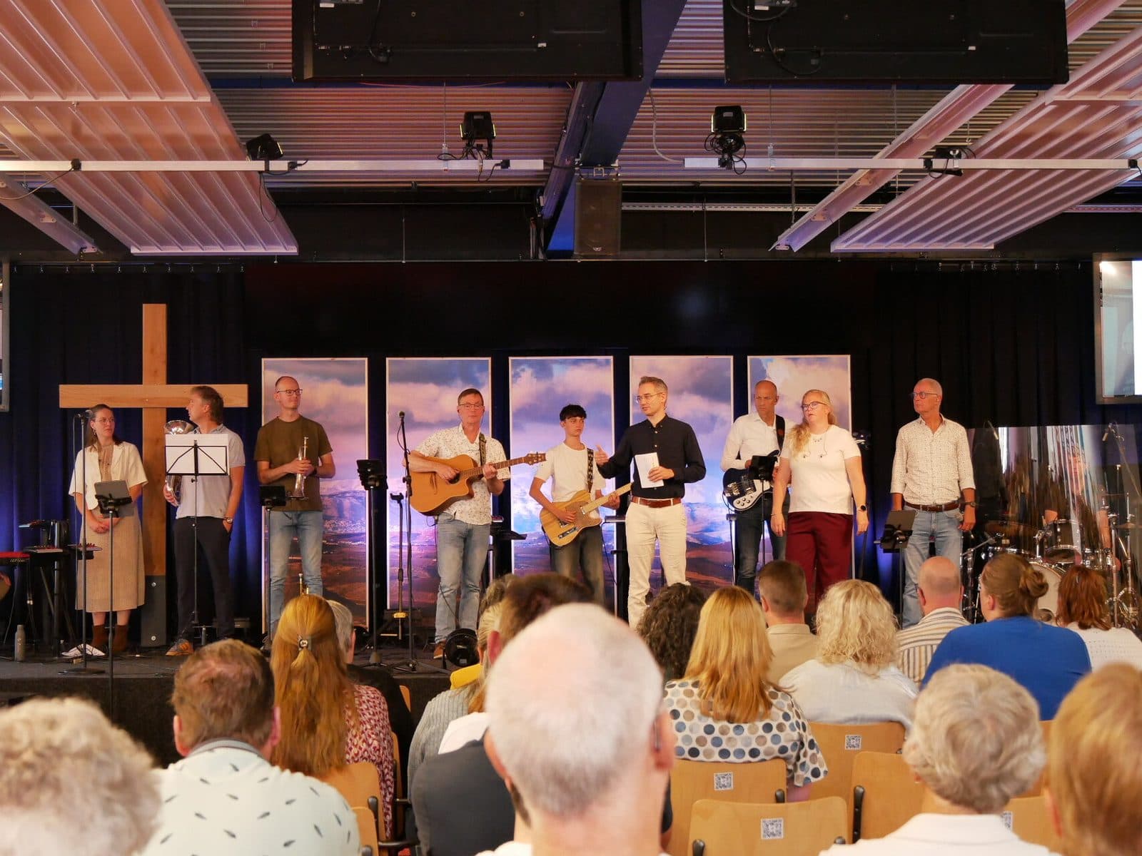 Het voltallige muziekteam op het podium naast het houten kruis: zang, trompet, gitaren, bas en drums, met de zaal ervoor.